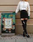 Model wearing ACQUO of Sweden over-the-knee glowing black boots, paired with a black mini skirt, cream bomber jacket, and teal gloves – striking urban fashion captured against a beige wall and colorful street art panel