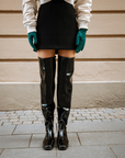 Front view of ACQUO of Sweden’s glowing black over-the-knee rubber boots paired with a black mini skirt, ivory bomber jacket, and emerald green gloves—Scandinavian luxury boots designed for bold, fashion-forward expression