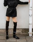 Model wearing ACQUO of Sweden over-the-knee glowing black boots with a flat sole, paired with a short black utility-style jacket – a bold, modern look set against a minimalist urban background