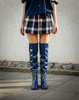 Front view of a model standing outdoors wearing Greta Iris Blue boots by ACQUO of Sweden. The image features iris blue over-the-knee rubber boots with a sleek reflective finish, styled with a navy shirt and plaid mini skirt against a textured concrete wall and cobblestone pavement.