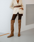 Woman wearing Greta Walnut Brown knee-high boots by ACQUO of Sweden styled with a cream belted blazer, dark shorts and tights in a minimalist indoor setting. Elegant waterproof boots with refined Swedish design and a modern fashion silhouette