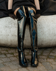 Model sitting outdoors wearing ACQUO of Sweden over-the-knee black natural rubber boots styled with an elegant black dress
