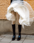 Woman wearing knee-high black rubber boots from ACQUO of Sweden paired with patterned white tights and a flowing layered white skirt, captured in motion against a rustic stone wall—an elegant fusion of vintage and modern Scandinavian style