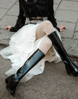 Model wearing ACQUO of Sweden knee-high glowing black boots with block heel, styled with a white layered tulle skirt, patterned white tights, and a black long-sleeve top – a bold and elegant street style look