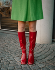 Model wearing Karin Cherry Red knee-high boots by ACQUO of Sweden styled with a green coat on a cobblestone street. Waterproof boots featuring elegant Swedish design and timeless Scandinavian fashion