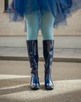 Front view of Karin Iris Blue knee-high rubber boots by ACQUO of Sweden styled with light blue tights and a navy tulle skirt outdoors. The elegant blue waterproof boots feature a sleek reflective finish and contemporary Scandinavian design.