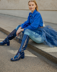 Model sitting outdoors in Karin Iris Blue knee-high rubber boots by ACQUO of Sweden, styled with a cobalt blue blouse, navy tulle skirt and light blue tights. The elegant blue waterproof boots feature a sleek reflective finish and timeless Swedish design