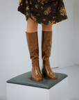 Close-up of Karin Walnut Brown knee-high boots by ACQUO of Sweden styled with a floral dress while standing on a pedestal in a minimalist studio. Elegant waterproof boots with refined Swedish design, square toes and a modern feminine silhouette