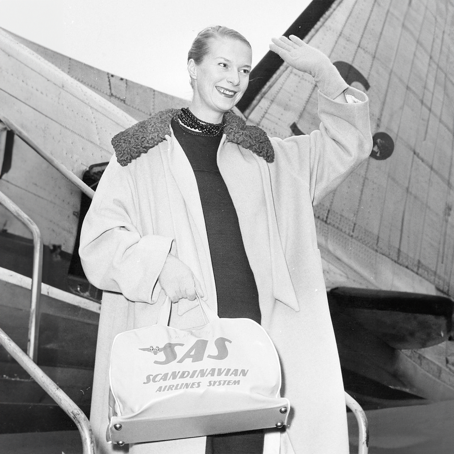 Katja of Sweden stepping off an airplane, dressed in a long elegant coat and holding a classic SAS travel bag