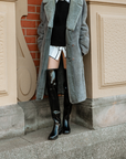 Model wearing ACQUO of Sweden over-the-knee glowing black boots, styled with a white shirt dress, black knit vest, and long grey shearling coat – a chic layered look combining modern fashion with classic Scandinavian outerwear