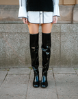 Close-up of model wearing ACQUO of Sweden over-the-knee glowing black boots, styled with a white shirt dress and black knitted vest – modern, statement footwear with a Scandinavian street style edge