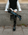 Model wearing ACQUO of Sweden over-the-knee glowing black boots, styled with a white oversized shirt, black knitted vest, and relaxed pose against a textured urban wall – a bold and modern Scandinavian fashion look