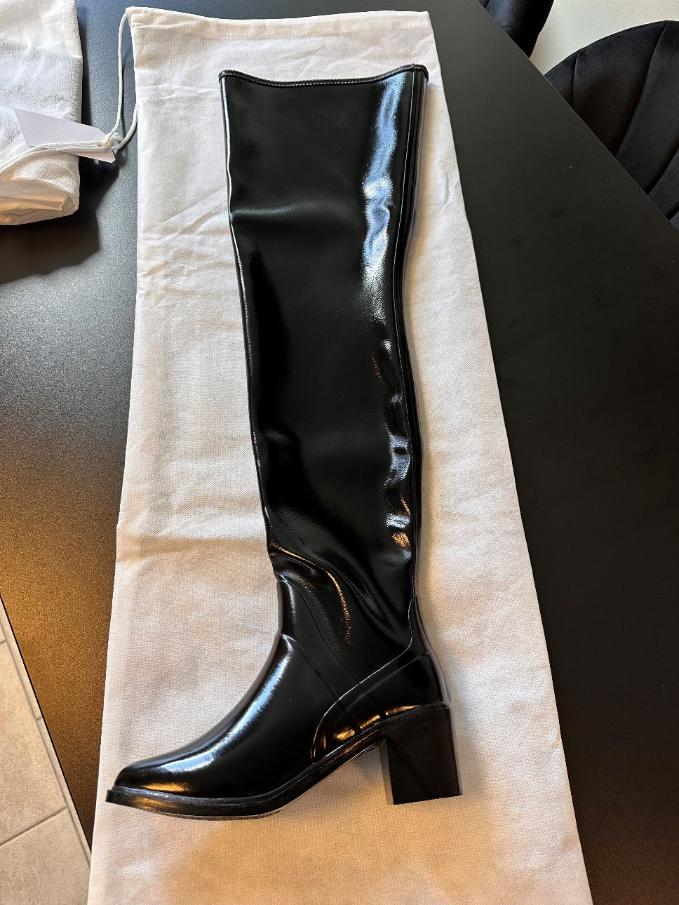 ACQUO of Sweden's signature black high-shine over-the-knee rubber boot with block heel, displayed on a white dust bag atop a black designer table — showcasing premium craftsmanship and Swedish fashion elegance