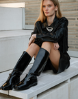 Model in a trendy black leather jacket with fringes and sleek shine knee-high boots posing seated on a white concrete staircase in front of a modern brick wall – minimalist street style look