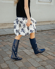 Side view of a model walking on a cobblestone street wearing Svea Iris Blue boots by ACQUO of Sweden. The image features iris blue knee-high rubber boots with a sleek reflective finish, styled with a black top and white floral skirt in an urban outdoor setting