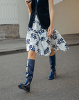 Model walking outdoors in Svea Iris Blue knee-high rubber boots by ACQUO of Sweden, styled with a white floral skirt and navy cardigan. The image highlights elegant blue waterproof boots with a sleek reflective finish in a contemporary street-style setting