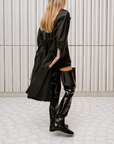 Model dressed in ACQUO of Sweden high-shine over-the-knee black boots, paired with a black embellished top, short bottoms, and a flowing black trench coat – a bold and elegant look set in a modern Scandinavian space