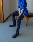 Model seated on a wooden bench wearing Vega Iris Blue boots by ACQUO of Sweden. The image highlights elegant iris blue over-the-knee rubber boots with a sleek fitted silhouette, square toe, and reflective finish, styled with a vibrant blue belted mini dress in a softly lit modern interior with wooden wall details.