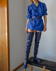 Model standing on a black leather bench wearing Vega Iris Blue boots by ACQUO of Sweden. The image features elegant iris blue over-the-knee rubber boots with a sleek fitted silhouette, square toe, and reflective finish, styled with a vibrant blue belted mini dress in a modern indoor setting with wooden details and natural light