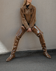 Woman wearing Vega Walnut Brown over-the-knee boots by ACQUO of Sweden paired with a brown jacket and black outfit against a textured wall. Elegant waterproof boots with a sleek Scandinavian design and modern fashion styling