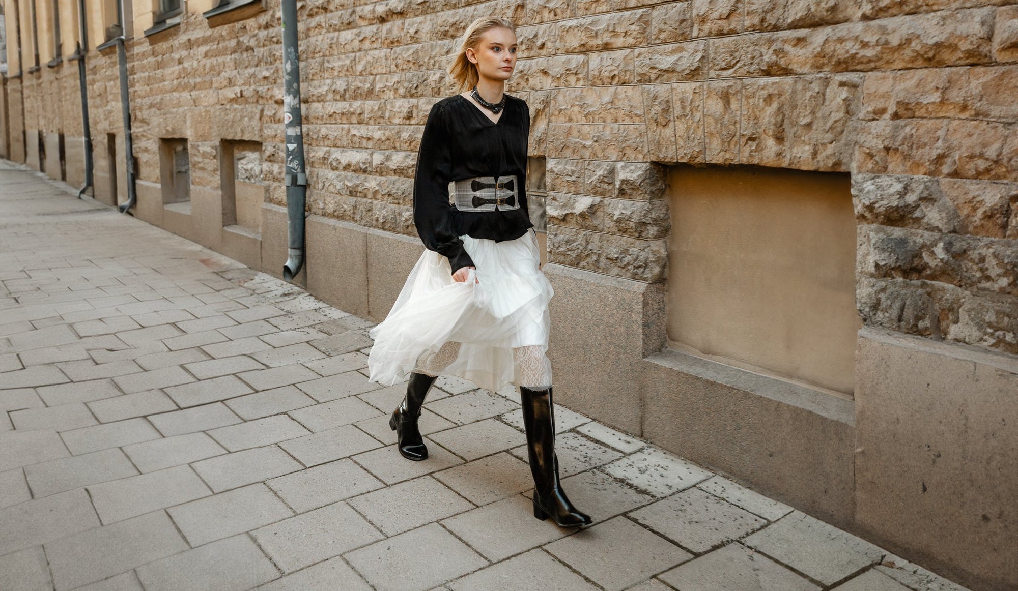 Model wearing ACQUO of Sweden knee-high glowing black heeled boots, styled with a voluminous white tulle skirt, lace-patterned tights, and a black blouse – a creative and elegant street style