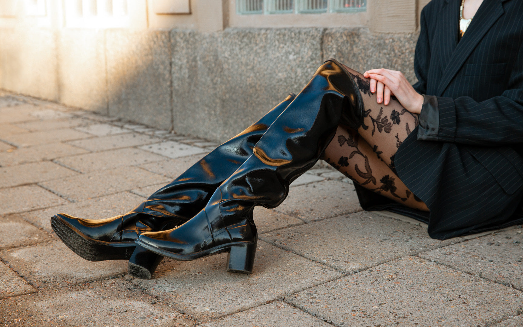 Model wearing ACQUO of Sweden knee-high glowing black heeled boots, styled with a black blazer, floral tights, and a statement necklace – a trendy and sophisticated city look in an urban setting