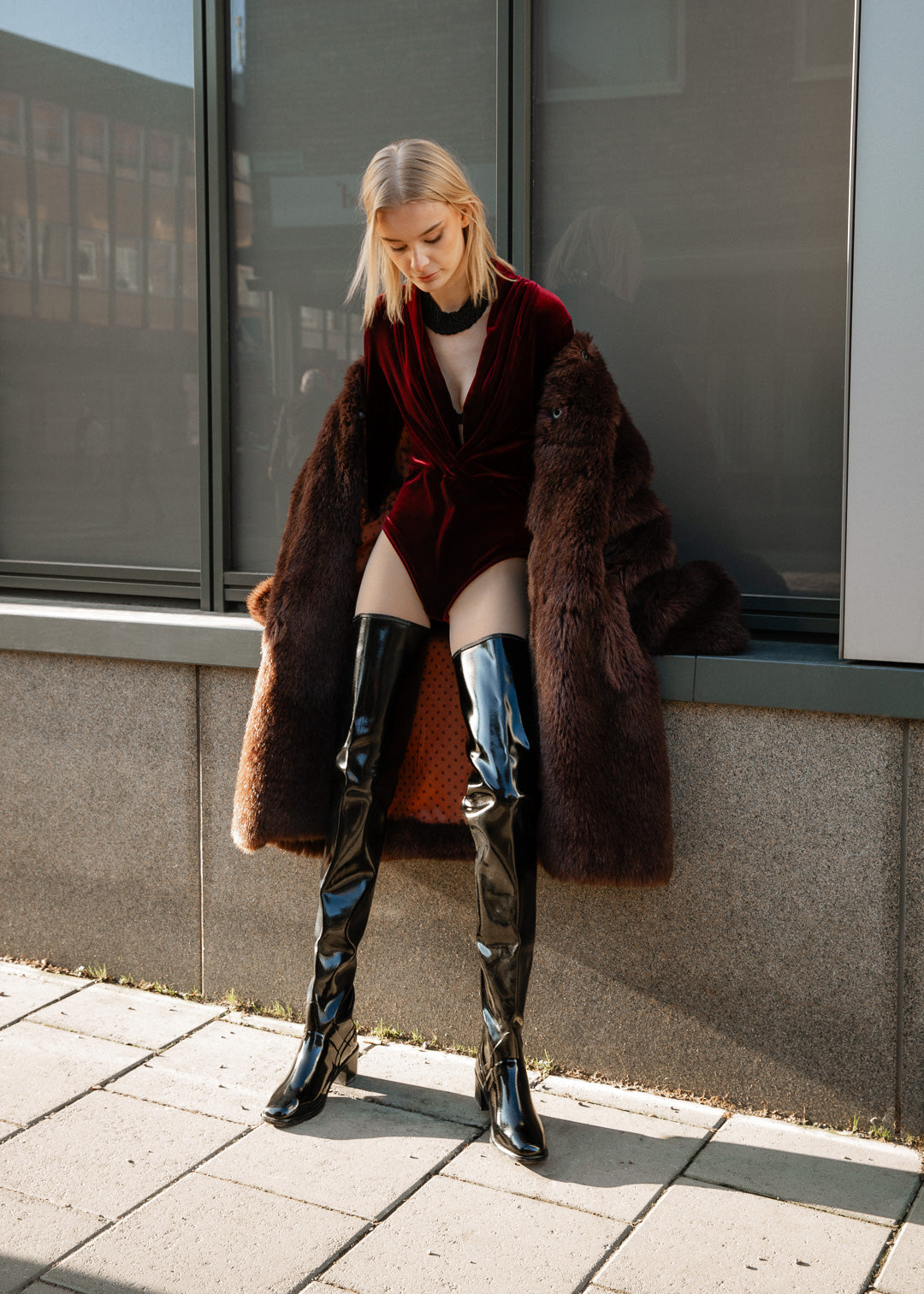 Fashion-forward model wearing ACQUO of Sweden over-the-knee glossy black rubber boots styled with a deep red velvet bodysuit and luxurious coat, showcasing a bold and elegant street style look perfect for modern, confident women.