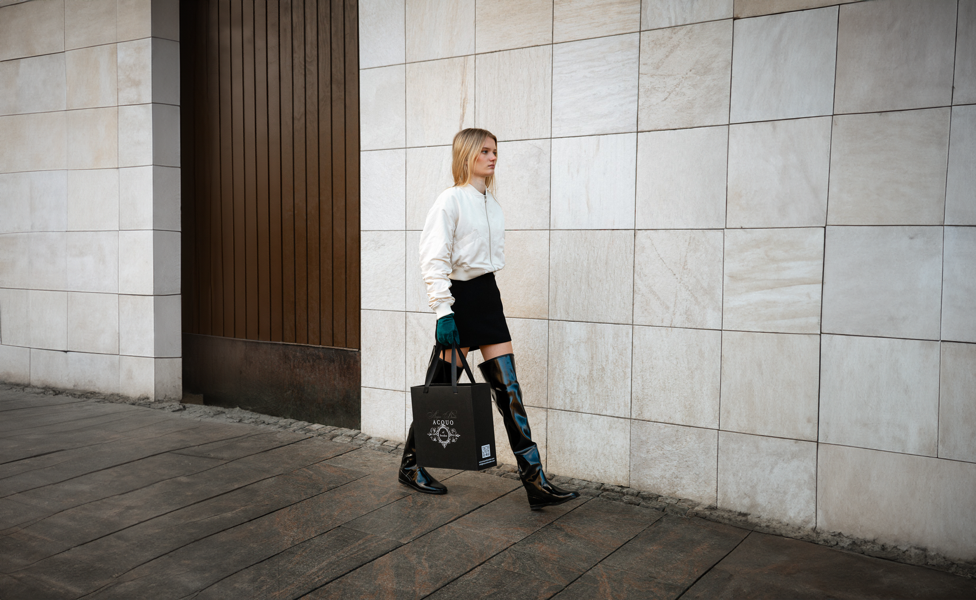 Stylish woman walking confidently along a stone wall in a chic urban outfit, wearing black over-the-knee ACQUO of Sweden boots, a white bomber jacket, and a black mini skirt, while holding an elegant black shopping bag with the ACQUO logo—embodying modern Swedish fashion with timeless elegance