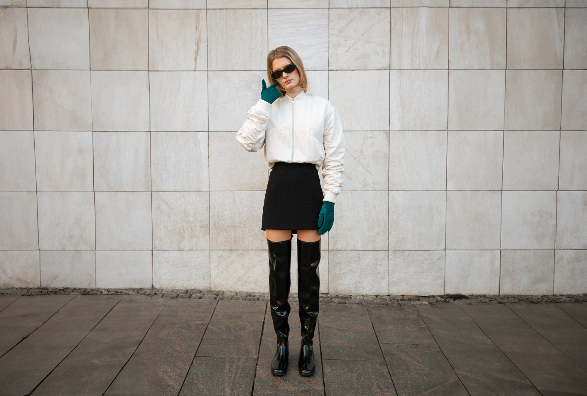 Fashion-forward woman in a monochrome outfit featuring black over-the-knee ACQUO of Sweden boots, a black mini skirt, white bomber jacket, dark sunglasses, and green gloves, posing confidently against a minimalist stone wall—highlighting modern Scandinavian style with bold, elegant footwear