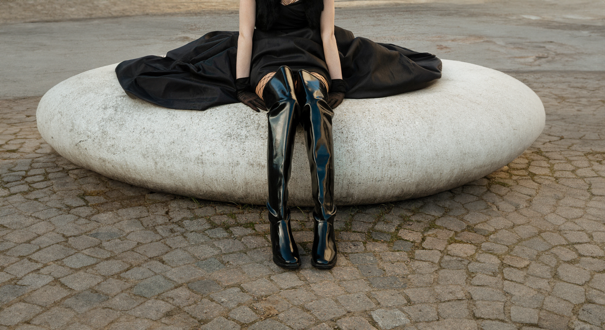 Model sitting outdoors wearing ACQUO of Sweden over-the-knee black natural rubber boots styled with an elegant black dress