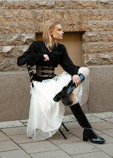 Stylish woman in ACQUO of Sweden's signature black the knee high rubber boots paired with a flowing white tulle skirt and structured black blouse, sitting confidently on a city street against a stone wall, showcasing bold Swedish fashion and premium boot design.