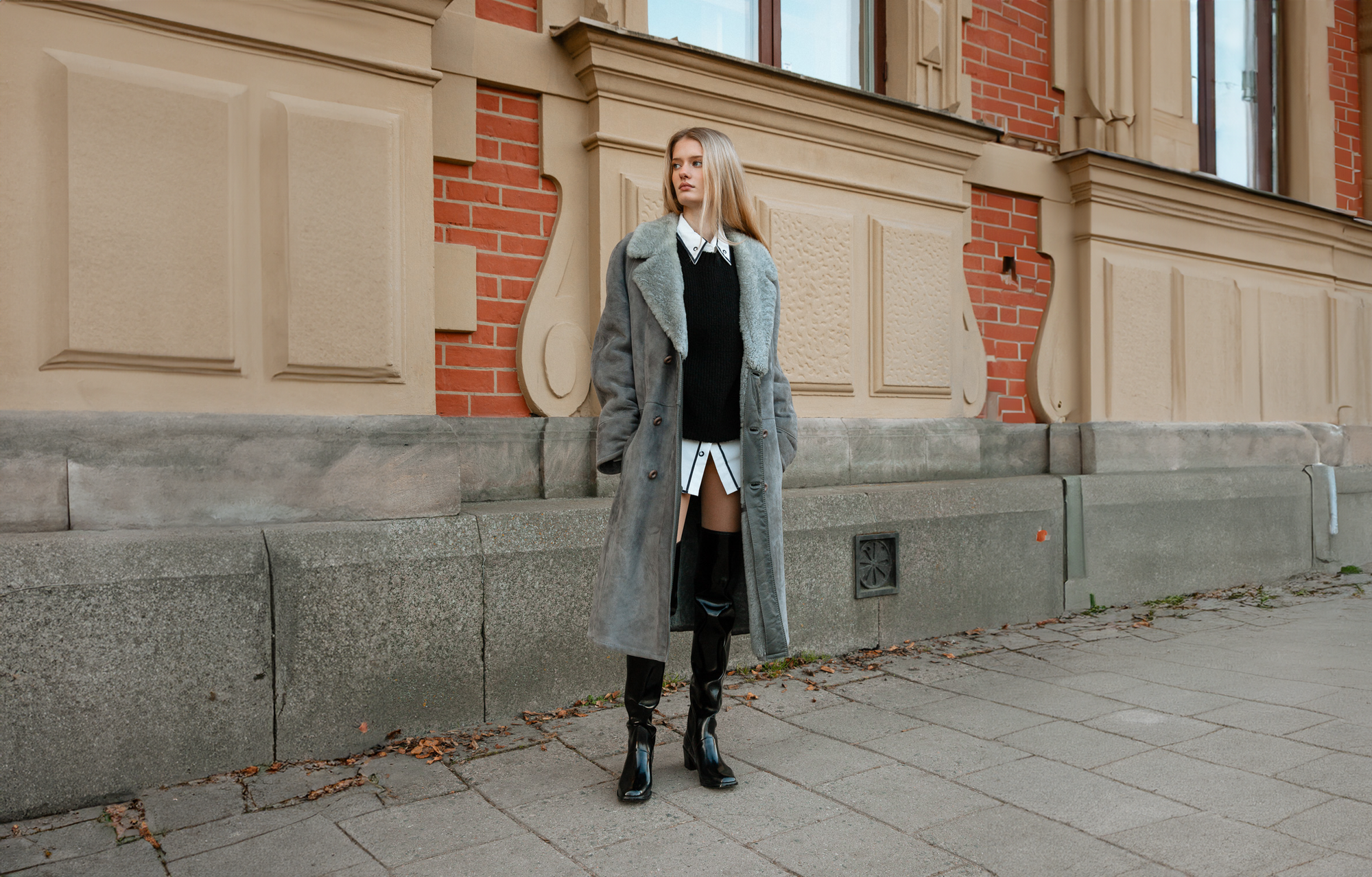 Fashionable woman standing in front of a historic building wearing ACQUO of Sweden over the knee black boots, a long grey shearling coat, and a layered black and white outfit — a sophisticated autumn street style look that highlights the bold, elegant design of ACQUO boots