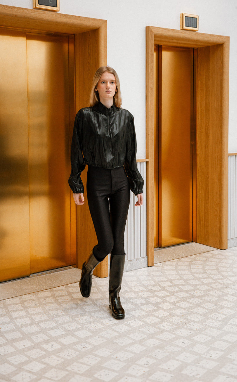 Elegant woman walking confidently in glossy black knee-high ACQUO boots, paired with high-waisted black pants and a silky black blouse, in front of luxurious golden elevators – showcasing Swedish fashion design by ACQUO of Sweden.