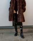 Model wearing ACQUO of Sweden over-the-knee glowing black heeled boots, styled with a deep red velvet bodysuit and oversized brown faux fur coat – a bold, luxurious street style look with Scandinavian elegance