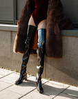 Model wearing ACQUO of Sweden's signature high-shine black over-the-knee boots with heels, styled with a deep red velvet bodysuit and luxurious brown faux coat – an elegant, bold fashion statement combining retro glamour and Scandinavian edge
