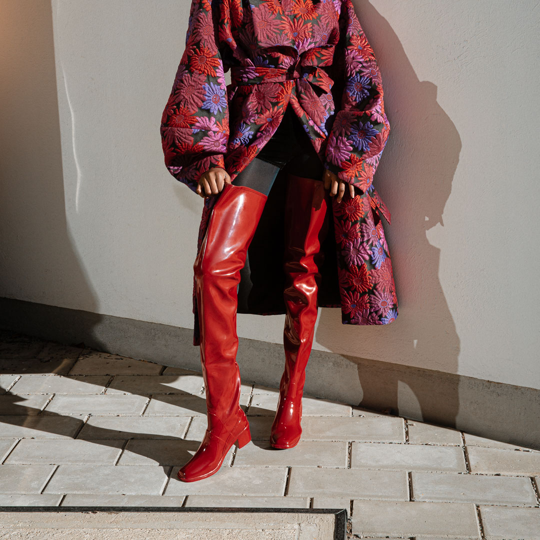 Model wearing Elsa Cherry Red over-the-knee rubber boots by ACQUO of Sweden, styled with a floral statement coat and black tights. Elegant waterproof boots with a bold silhouette inspired by contemporary Swedish design.