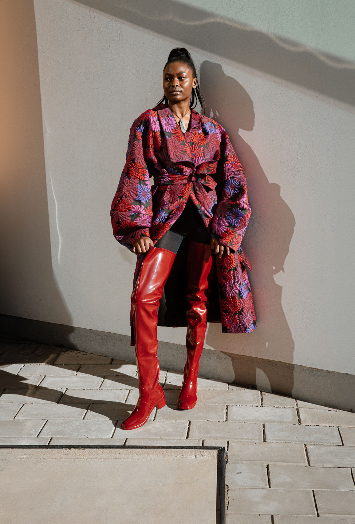 Model wearing Elsa Cherry Red over-the-knee rubber boots by ACQUO of Sweden, styled with a dramatic floral coat and black tights. Elegant waterproof fashion boots with a bold silhouette inspired by modern Swedish design.