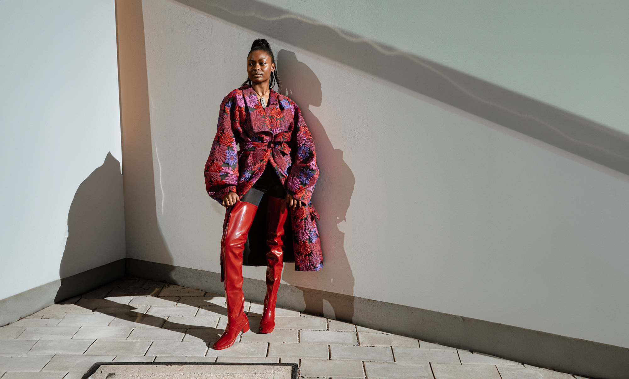 Model wearing Elsa Cherry Red over-the-knee rubber boots by ACQUO of Sweden, styled with a dramatic floral coat and black tights. Elegant waterproof fashion boots with a bold silhouette inspired by modern Swedish design.