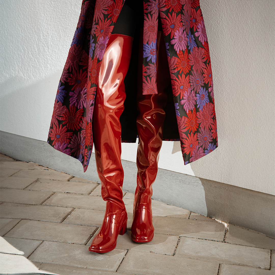Close-up of Elsa Cherry Red over-the-knee rubber boots by ACQUO of Sweden, styled with a floral jacquard coat and black tights. Bold waterproof boots with a sleek silhouette and refined Swedish design.