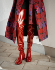 Close-up of Elsa Cherry Red over-the-knee rubber boots by ACQUO of Sweden, styled with a floral jacquard coat and black tights. Bold waterproof boots with a sleek silhouette and refined Swedish design.
