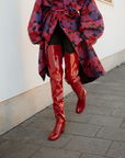 Model walking in Elsa Cherry Red over-the-knee rubber boots by ACQUO of Sweden, paired with black tights and a floral statement coat. Waterproof fashion boots with an elegant silhouette and contemporary Swedish design.