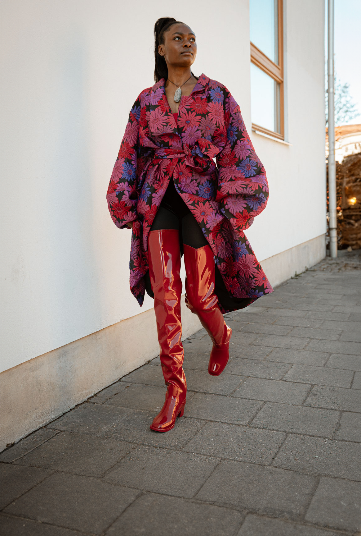 Model walking in Elsa Cherry Red over-the-knee rubber boots by ACQUO of Sweden, paired with a floral oversized coat and black tights. Statement waterproof boots with a sleek silhouette and contemporary Swedish design