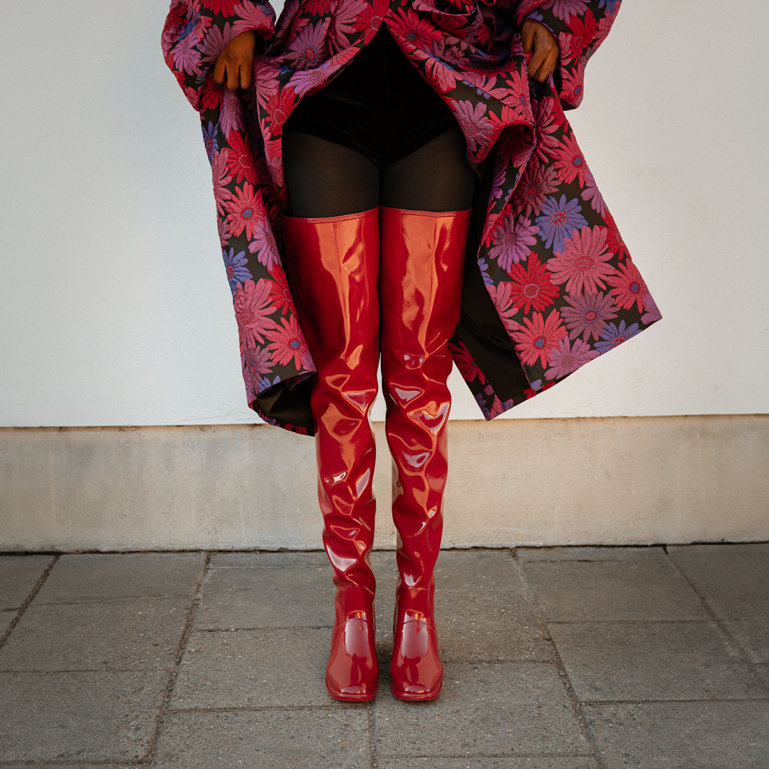 Front view of Elsa Cherry Red over-the-knee rubber boots by ACQUO of Sweden, styled with black tights and a floral coat. Statement waterproof boots with a sleek high-leg silhouette and elegant Swedish design