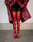 Front view of Elsa Cherry Red over-the-knee rubber boots by ACQUO of Sweden, styled with black tights and a floral coat. Statement waterproof boots with a sleek high-leg silhouette and elegant Swedish design