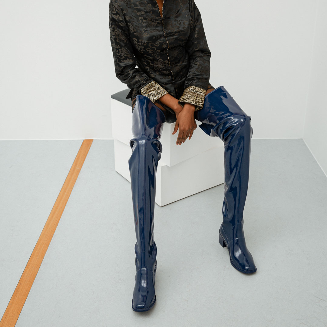 Model seated on white display cubes wearing Elsa Iris Blue boots by ACQUO of Sweden. The image shows tall iris blue rubber boots with a sleek fitted silhouette, square toe, and low block heel, styled with a dark patterned jacket in a minimalist studio setting