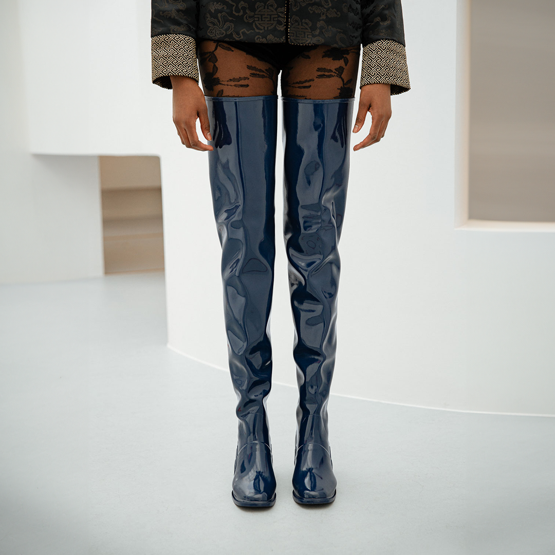 Front view of a model wearing Elsa Iris Blue boots by ACQUO of Sweden in a minimalist studio interior. The image highlights tall iris blue rubber boots reaching above the knees, featuring a sleek fitted design, square toe, and reflective finish, styled with patterned black tights and a dark embroidered jacket