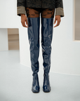 Front view of a model wearing Elsa Iris Blue boots by ACQUO of Sweden in a minimalist studio interior. The image highlights tall iris blue rubber boots reaching above the knees, featuring a sleek fitted design, square toe, and reflective finish, styled with patterned black tights and a dark embroidered jacket