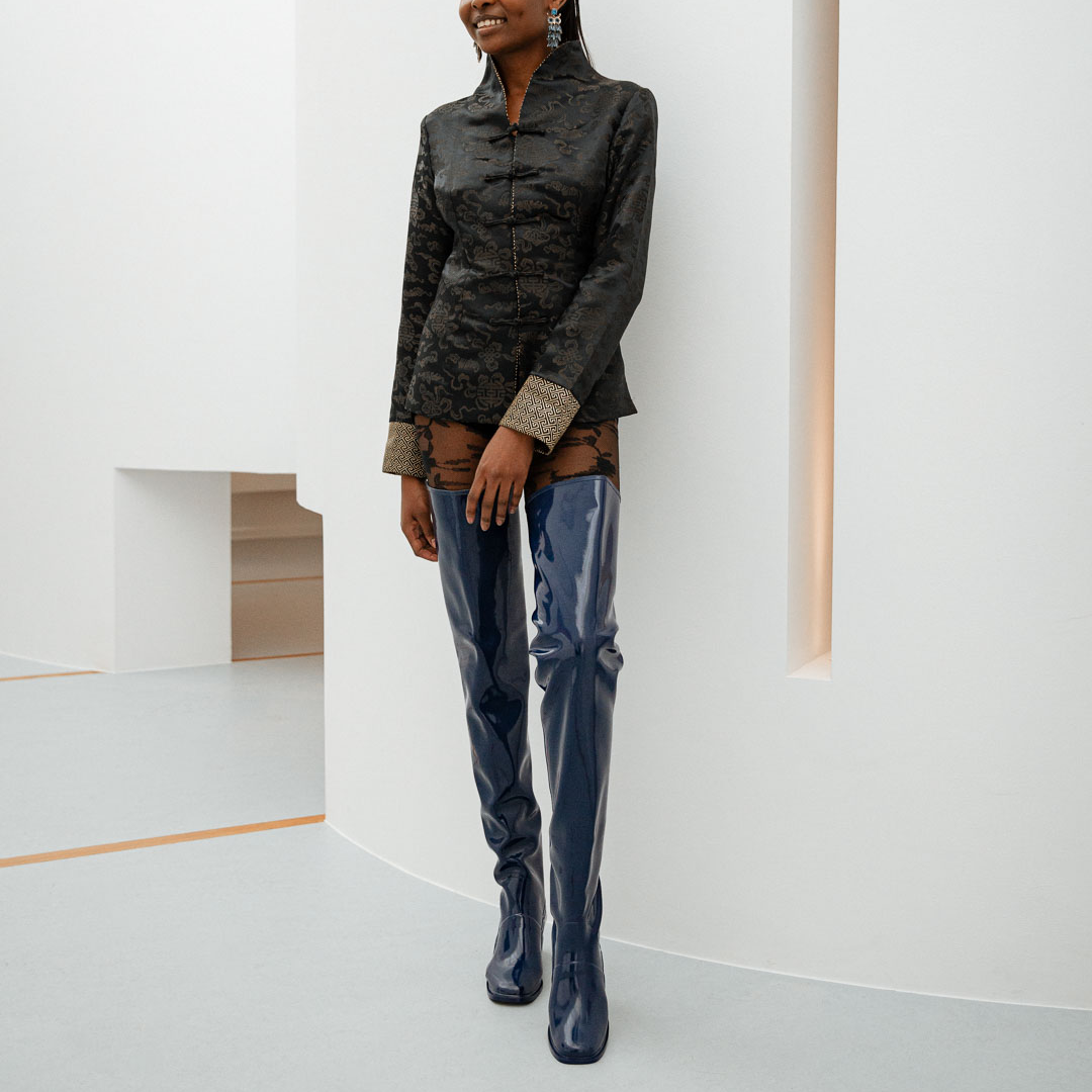 Model leaning against a white wall wearing Elsa Iris Blue boots by ACQUO of Sweden. The image shows elegant iris blue over-the-knee rubber boots with a sleek fitted silhouette, square toe, and reflective finish, styled with patterned tights and a tailored black jacquard jacket in a minimalist studio interior.