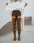 Woman wearing Elsa Walnut Brown over-the-knee boots by ACQUO of Sweden with a white textured top and bronze sequined shorts in a modern indoor setting. Waterproof boots featuring elegant Swedish design and contemporary Scandinavian fashion