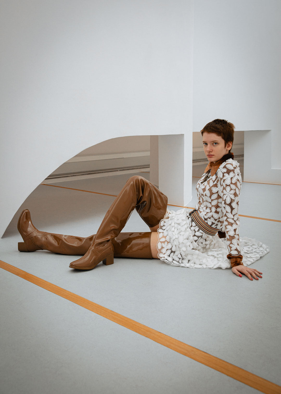Woman sitting on the floor in a modern interior wearing Elsa Walnut Brown over-the-knee boots by ACQUO of Sweden with a white floral lace dress. Waterproof boots featuring elegant Swedish design and refined Scandinavian fashion styling